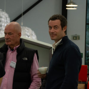 two attendees of an event in a room, one looks at the camera with a tea cup in his hand